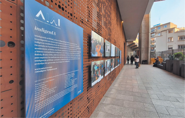A photograph of people looking at exhibition materials comprising an introductory panel entitled ‘AIAI’ and ‘indigenIA’, and a set of photographs of people undertaking a workshop. The exhibition is on the outside of a building and the images are attached to a metal wall with lots of regular holes in it. Other buildings and blue sky can be seen in the distance.